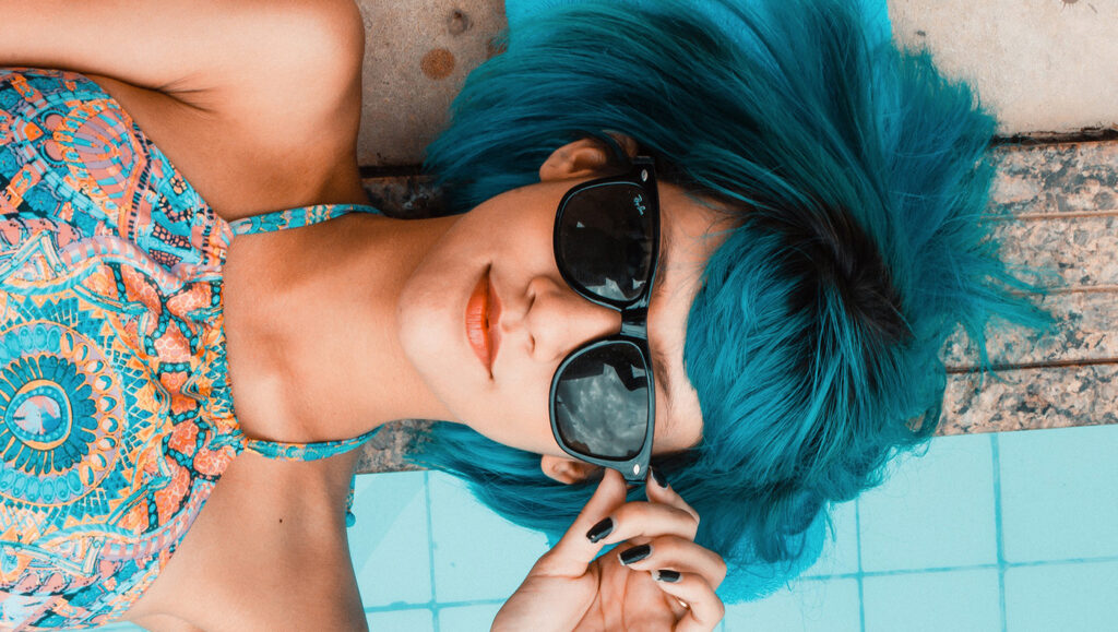 portrait d'une femme aux cheveux bleus avec des lunettes de soleils couchée sur le bord d'une piscine pendant son cycle menstruel